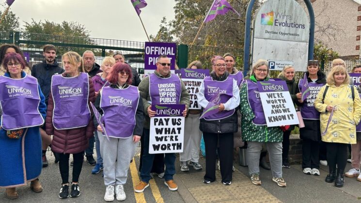 A group of people standing on a picket line wearing UNISON tabards and carrying banners stating "Pay us what we're worth".